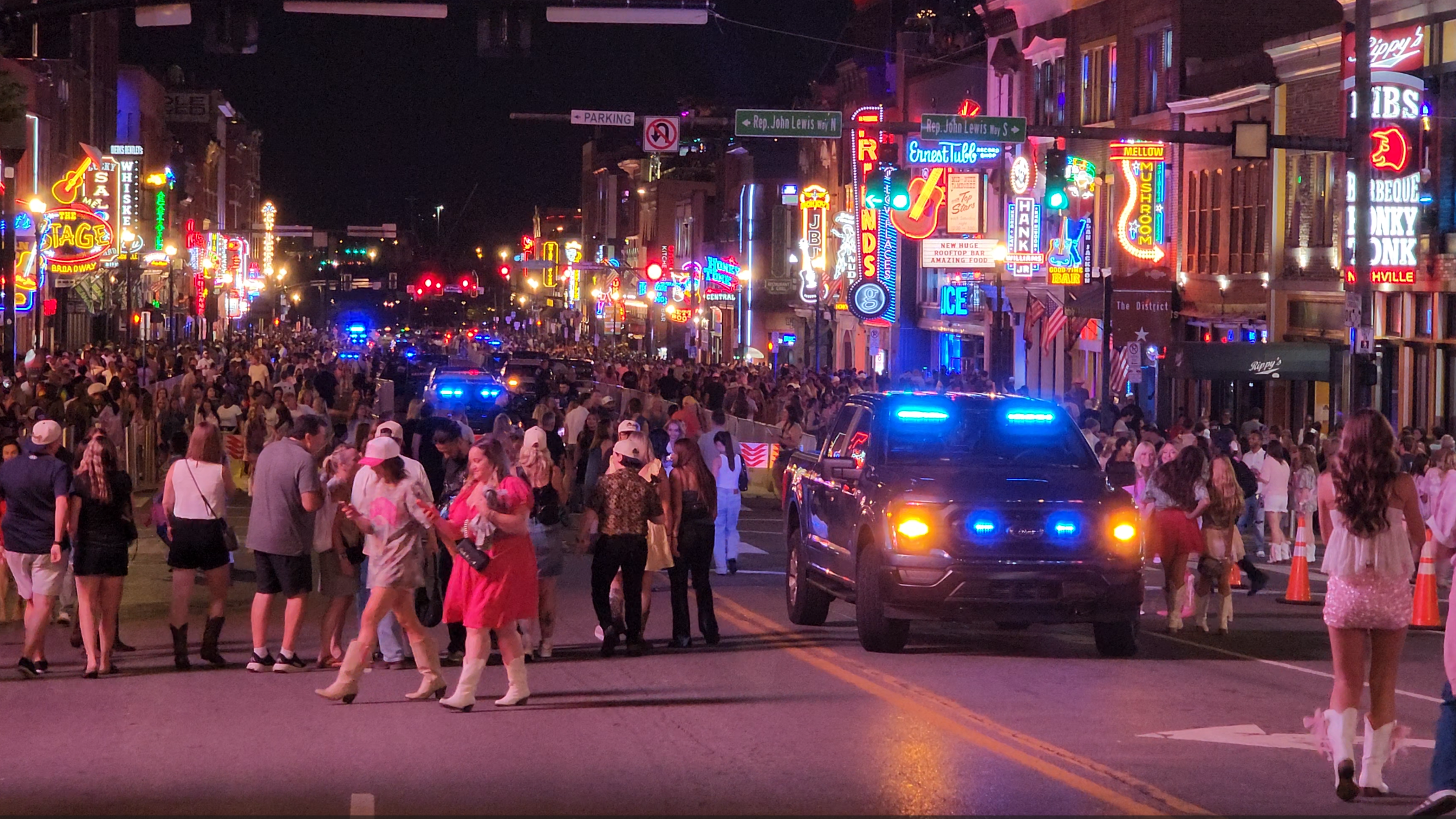 A large nighttime crowd filling Lower Broadway beneath neon honky-tonk signs as a police vehicle moves through the street.